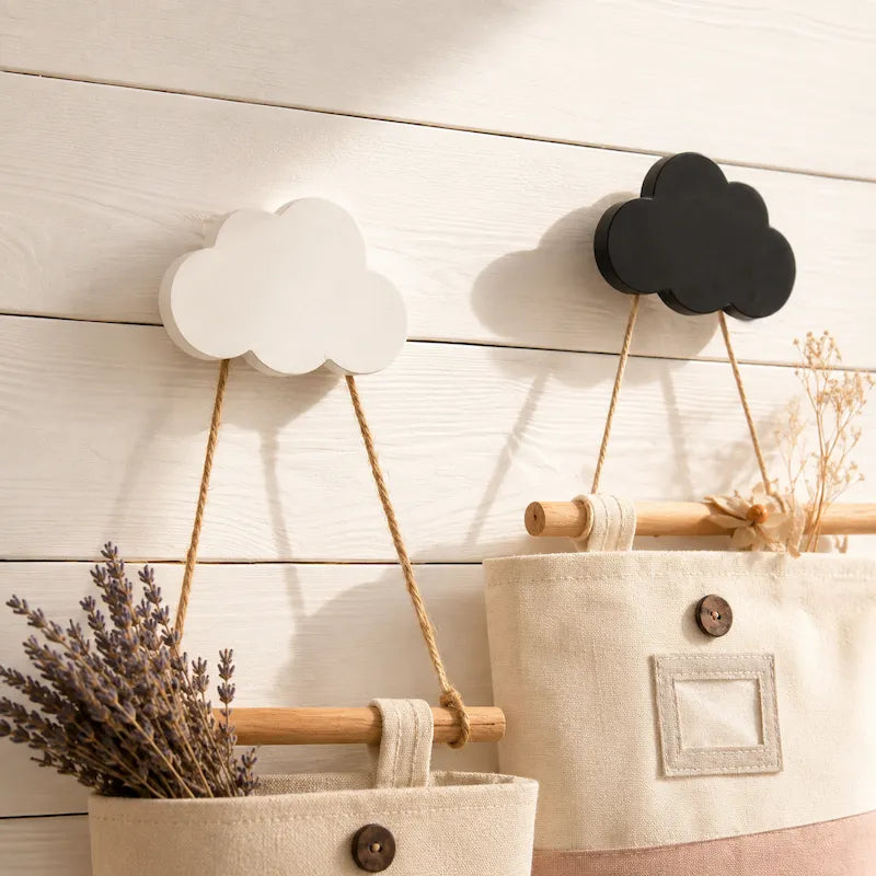 Two cloud-shaped wall hooks hang on a white wood plank wall. One hook is white and the other is black, each suspended by rope from a wooden rod with a canvas hanging basket below.