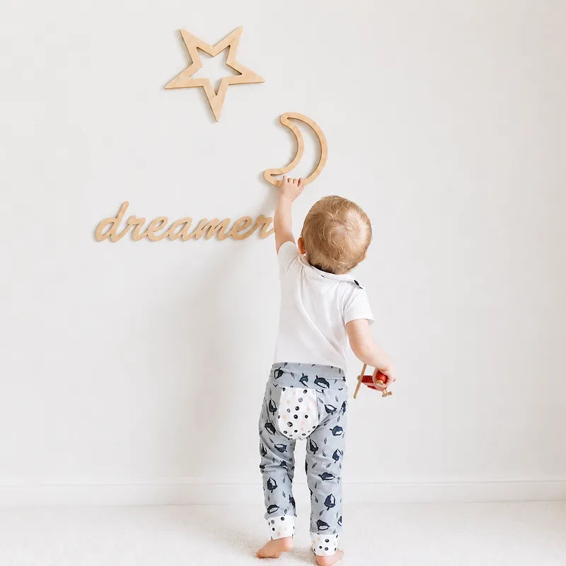 A toddler stands with their back to the camera, reaching for a wooden wall decoration. The room features a white wall decorated with a wooden star, a wooden crescent moon, and the word 'dreamer' in cursive.
