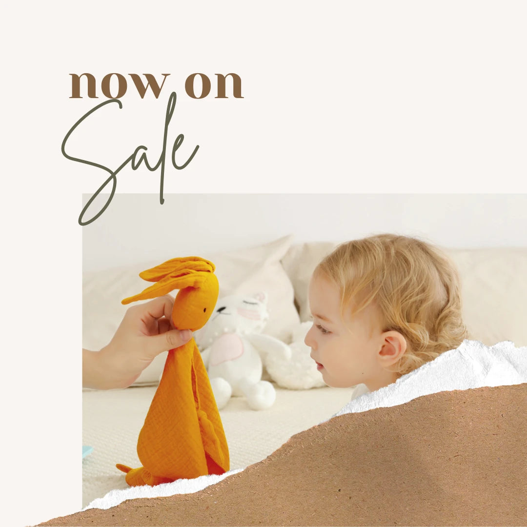 A baby or toddler lying under a white blanket on a bed, looking at an orange fabric bunny toy held by an adult hand. A white plush toy is behind them, and the top-left corner has the text 'now on Sale' on a cream background.