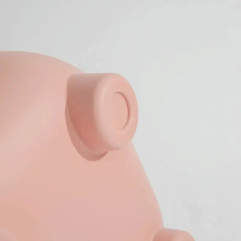 Close-up detail of a matte pink pig-shaped object, showing part of its rounded body and a small protruding circular feature against a very light gray background.