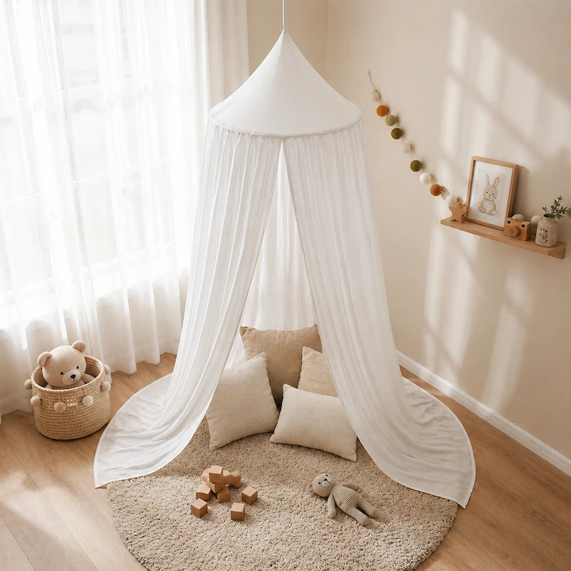 A white cotton canopy baby play tent draped over a round beige rug in a bright, sunlit nursery. The setup includes neutral-toned pillows, wooden blocks, a plush toy, a woven basket with a teddy bear, and a small wall shelf with a framed picture and decorative garland.